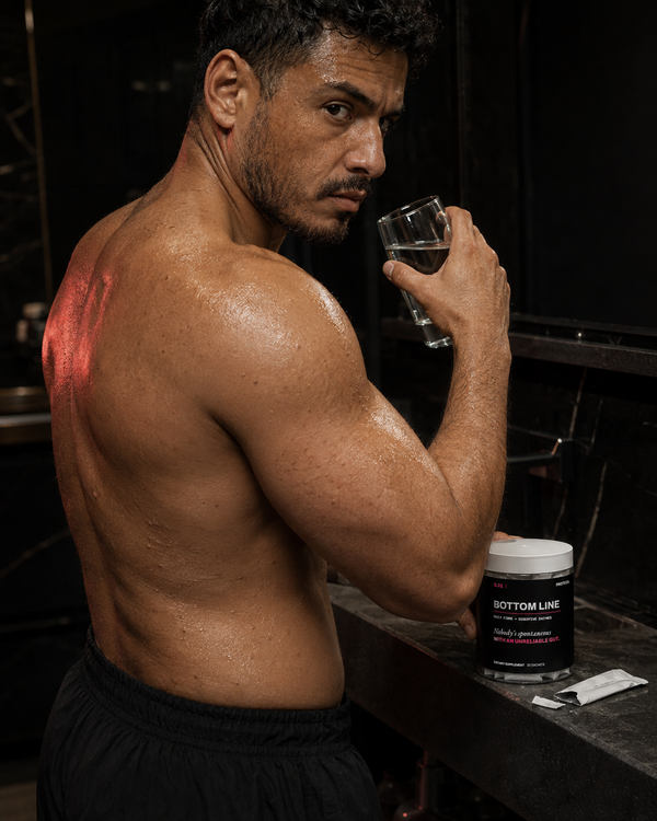 A shirtless man at a bar holding a glass under red light.