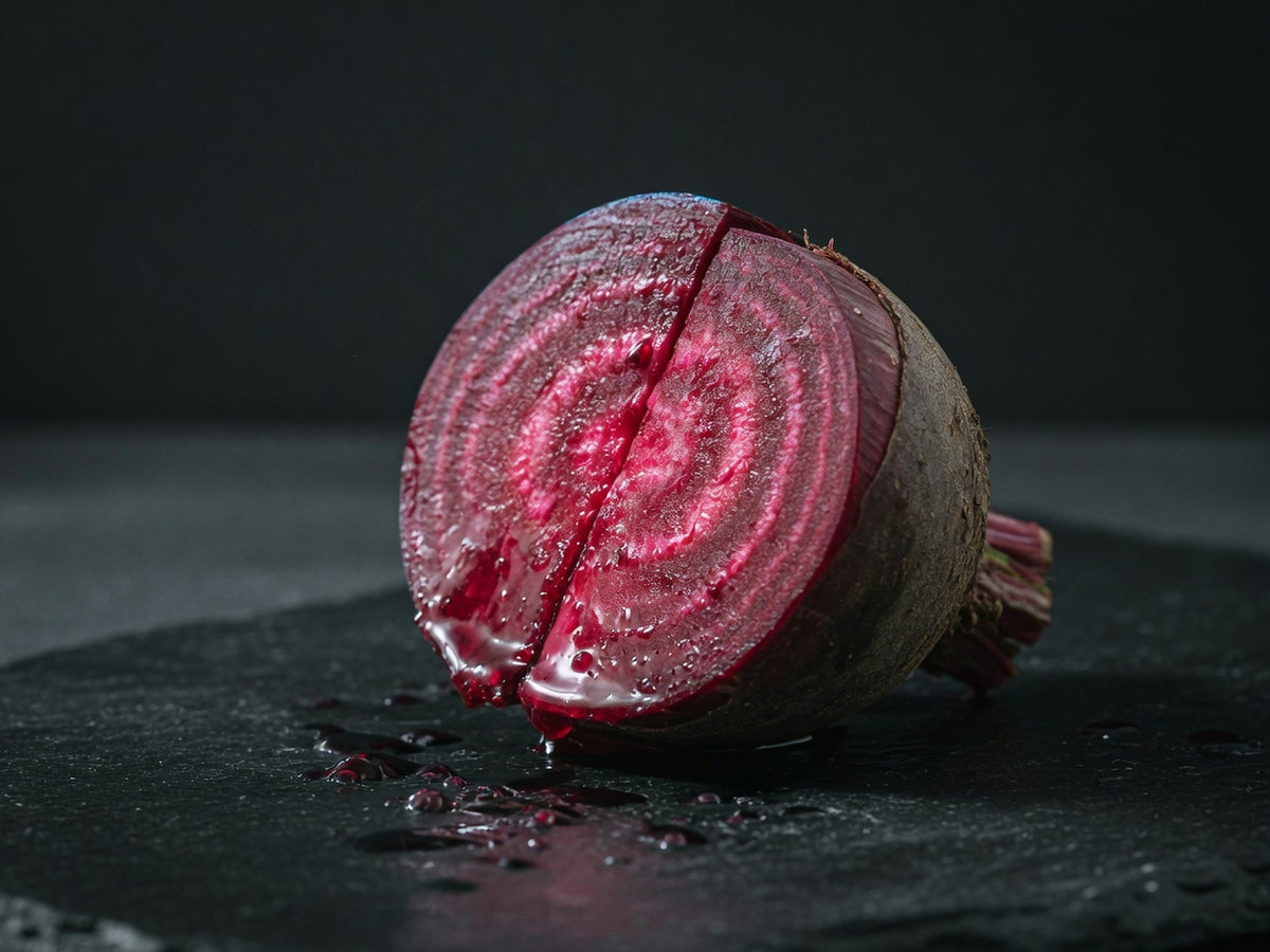 A raw beetroot cut in half, showing deep magenta concentric rings and wet earthy skin.