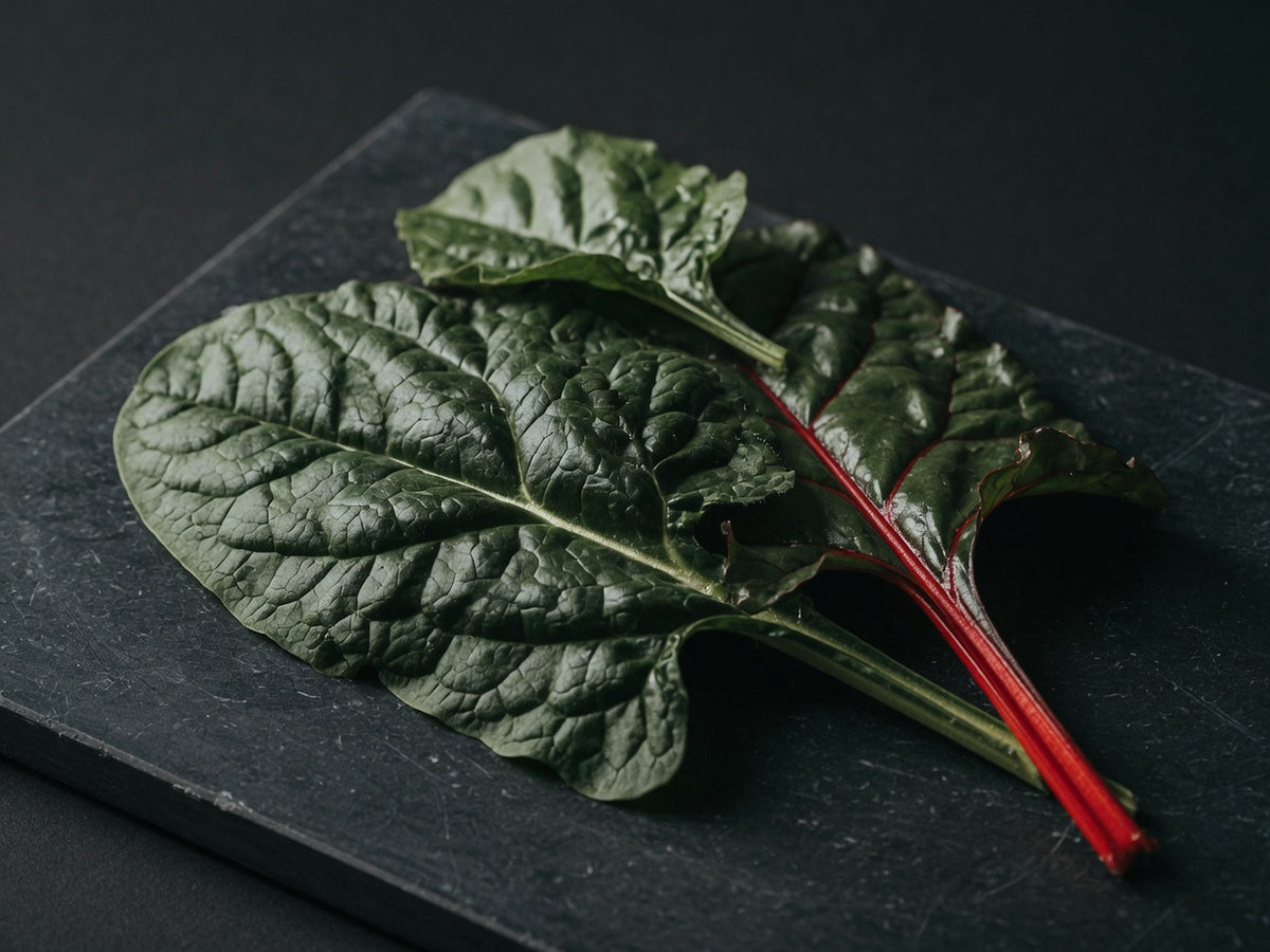 Dark green spinach and chard leaves with a red stem, overlapping on black stone.