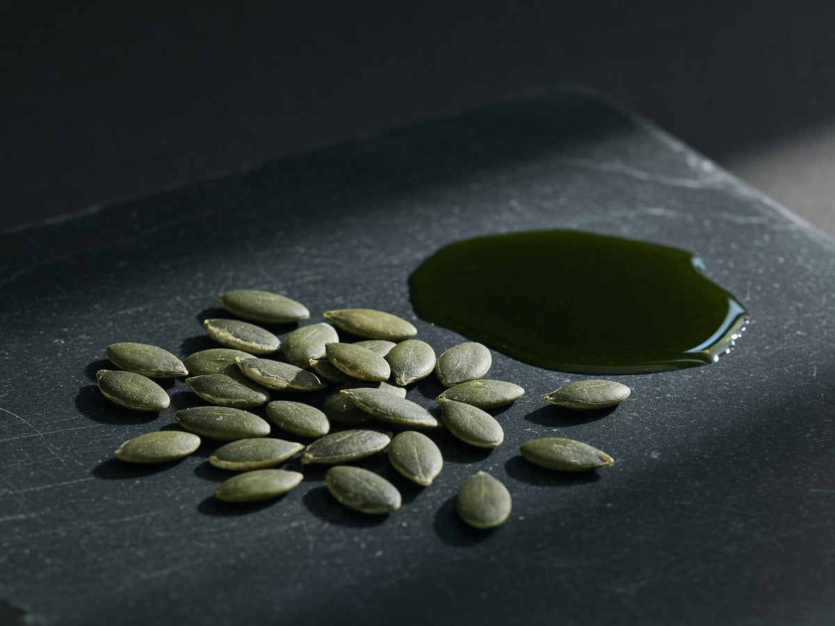 A scatter of flat green pumpkin seeds beside a pool of dark green pressed oil.