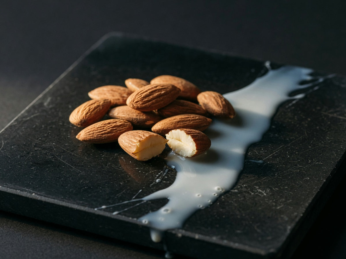 A pile of raw almonds in their brown skins beside a thin pour of milk on black stone.