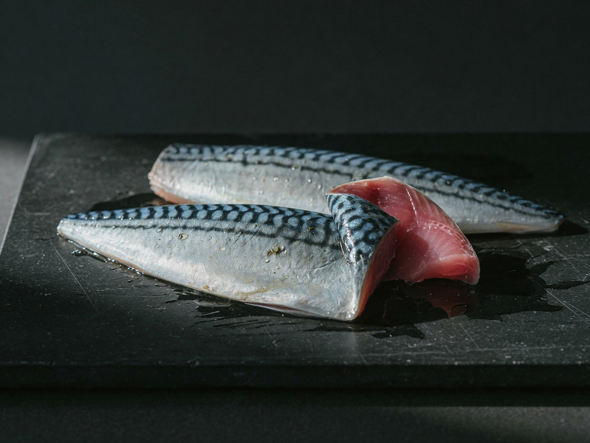 Two silver-blue mackerel fillets on black stone, one folded to show deep pink flesh.